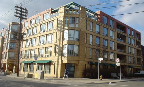 Five-storey building in buff brick occupies a corner lot, with green girders visible on a roof terrace